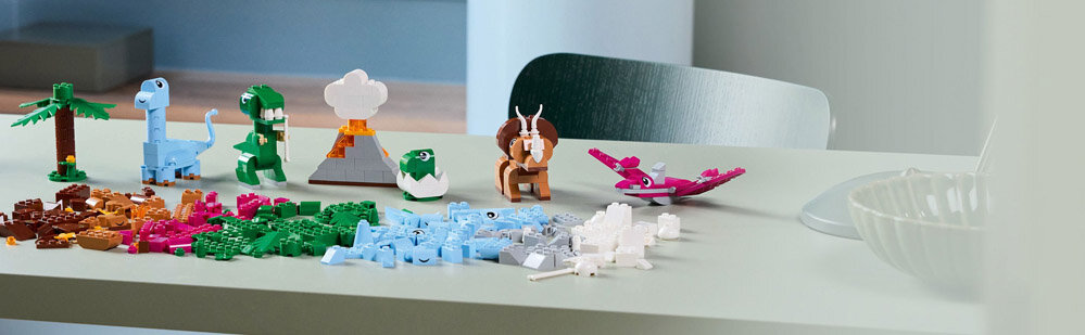 Na stole znajdują się różnokolorowe klocki LEGO ułożone w kształty przypominające dinozaury i inne obiekty, takie jak drzewo i wulkan. Obok nich leżą luźne klocki w różnych kolorach.