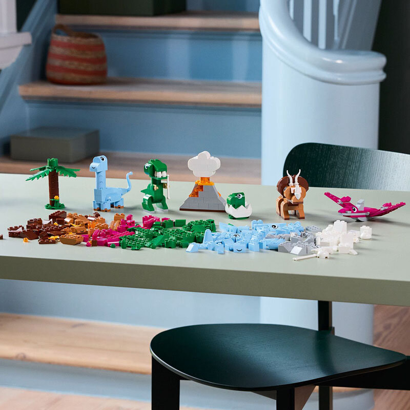 Na stole znajdują się różnokolorowe klocki LEGO ułożone w kształty przypominające dinozaury, wulkan i drzewo. W tle widoczne są schody i kosz.