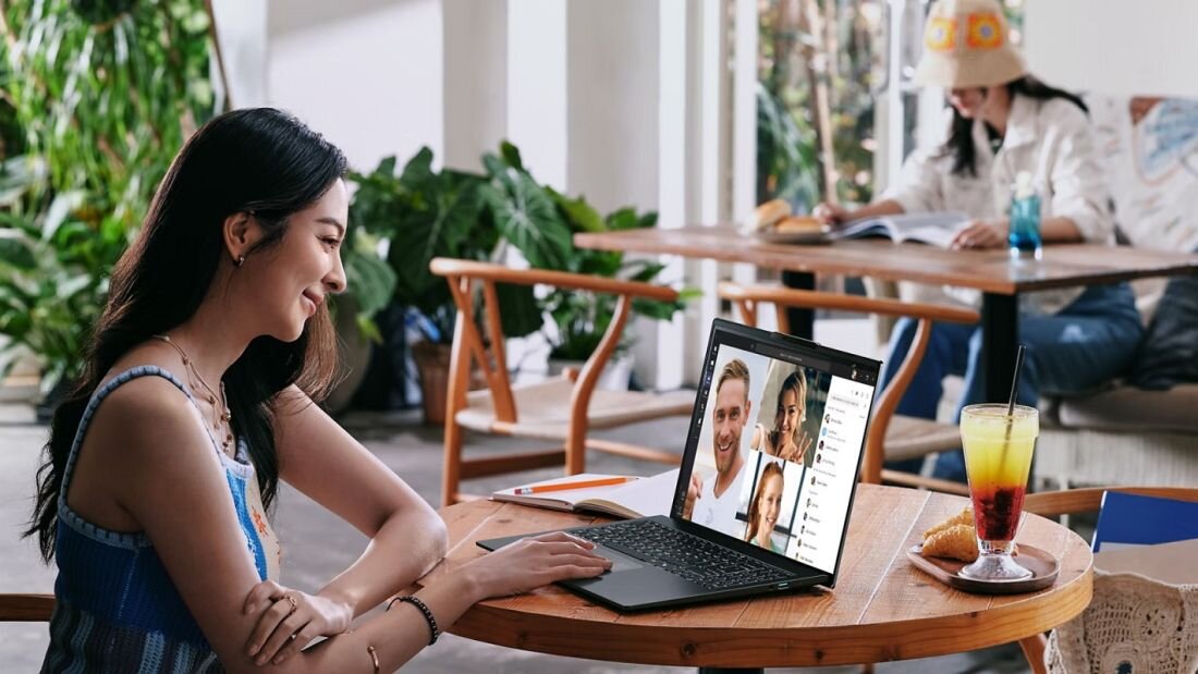 Kobieta w niebieskiej bluzie uśmiechająca się podczas wideokonferencji na laptopie Acer Swift Go 14 AI w kawiarni, na ekranie cztery osoby w oknach rozmowy wideo, obok kolorowy napój w szklance i rogalik na talerzu, w tle osoba w białej czapce, atmosfera pracy zdalnej z funkcją Wi-Fi 7. – Wi-Fi 7 i Bluetooth Kamera QHD IR