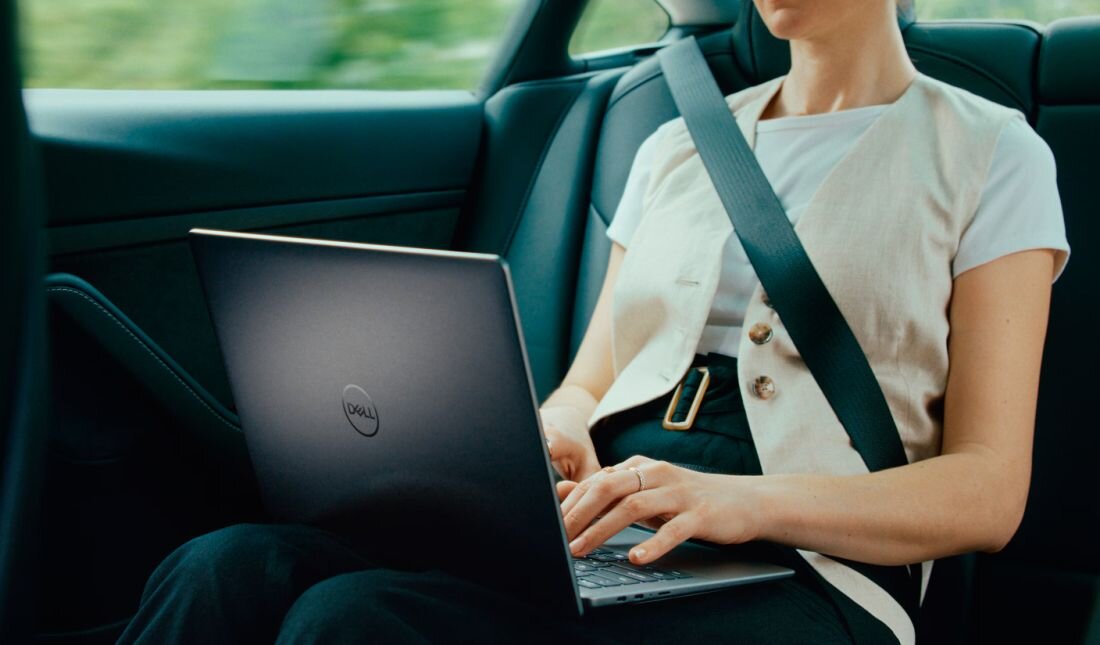 Kobieta w kamizelce podróżująca samochodem korzystająca z laptopa Dell Latitude na tylnym siedzeniu z zapiętym pasem bezpieczeństwa i zielonym tłem za oknem. - Qualcomm FastConnect Wi-Fi 7 