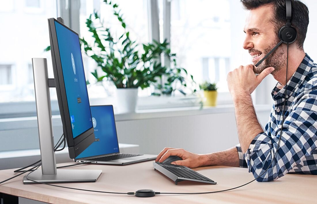 Uśmiechnięty mężczyzna w słuchawkach i kraciastej koszuli prowadzący wideokonferencję na zewnętrznym monitorze podłączonym do laptopa Dell w jasnym biurze. - LPDDR5X 8448 MHz Dysk SSD PCIe NVMe 4.0 