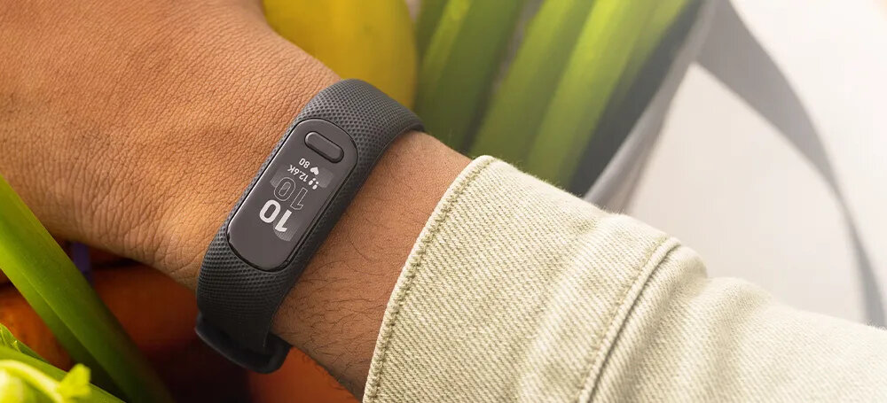 Na zdjęciu widoczny jest czarny smartband na nadgarstku osoby, z wyświetlaczem pokazującym liczbę 10 i symbolem baterii. W tle znajdują się zielone i żółte elementy, prawdopodobnie owoce lub warzywa.
