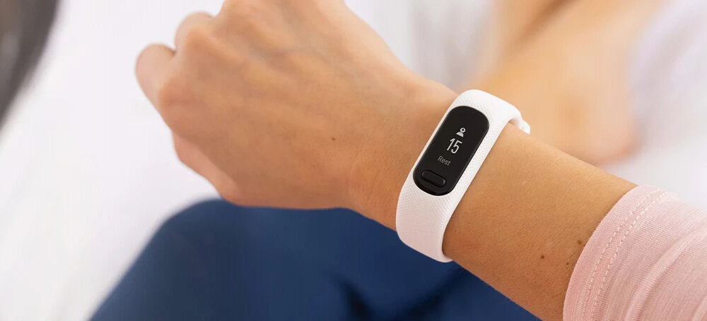 Osoba nosi biały smartband na nadgarstku, na ekranie widoczny jest numer 15 oraz napis \'Rest\'.