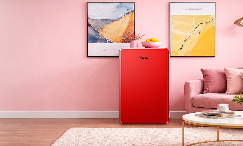 LODÓWKA MIDEA MDRD142SLE32 Zdjęcie przedstawia czerwoną lodówkę ustawioną w stylowym, różowym wnętrzu z obrazami na ścianie i sofą po prawej stronie. Na lodówce stoi misa z owocami, dodając kolorystycznego akcentu