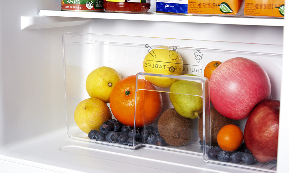 LODÓWKA MIDEA MDRD142SLE32 Na zdjęciu widać dolną szufladę lodówki wypełnioną świeżymi owocami takimi jak jabłka, cytrusy, gruszka, kiwi oraz borówki. Przezroczysta przednia ścianka pozwala łatwo zobaczyć zawartość 93 litrów pojemność przechowywanie żywności oszczędność miejsca kompaktowe wymiary