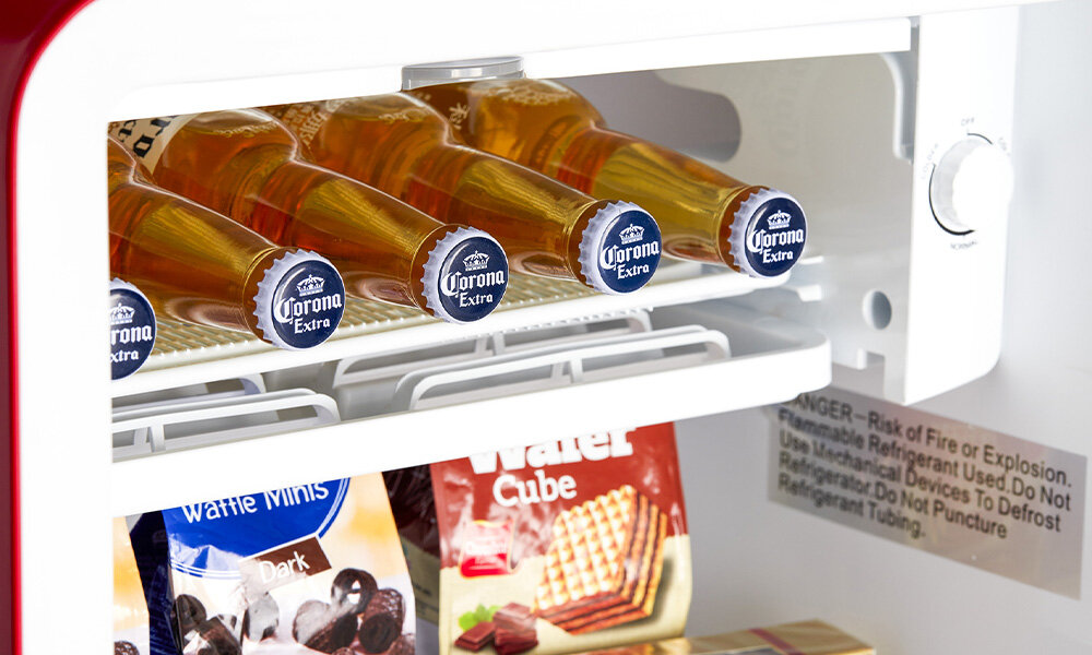 LODÓWKA MIDEA MDRD142SLE32 Zdjęcie pokazuje wnętrze lodówki, w której na górnej półce ułożono kilka butelek napoju w szklanych butelkach z etykietami skierowanymi w stronę aparatu. Niżej znajdują się paczkowane produkty, takie jak wafle komora niskich temperatur przechowywanie produktów chłodzenie napoje desery świeżość