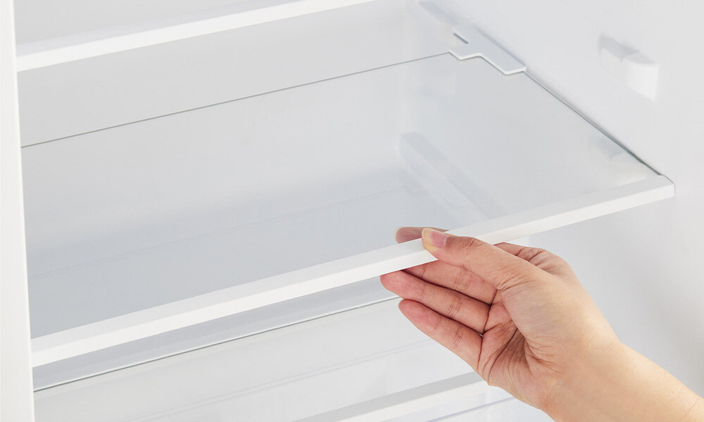 LODÓWKA MIDEA MDRD142SLE32 Zdjęcie przedstawia dłoń unoszącą szklaną półkę wewnątrz lodówki. Półka jest przezroczysta, a wnętrze urządzenia jasne i minimalistyczne szklana wysuwana półka pojemniki półki na drzwiach