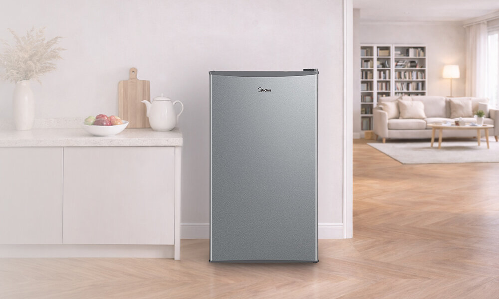 LODÓWKA MIDEA MDRD142FGE50 Zdjęcie przedstawia wolnostojącą, srebrną lodówkę ustawioną w urządzonej, jasnej przestrzeni. W tle widać blat kuchenny oraz otwarte przejście do salonu z kanapą i regałami