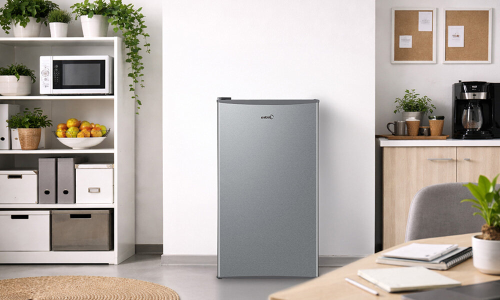 LODÓWKA MIDEA MDRD142FGE50 Na zdjęciu widać wolnostojącą, srebrną lodówkę ustawioną w jasnym pomieszczeniu z półkami, roślinami i sprzętami kuchennymi. Obok znajduje się biurko z notatnikiem, co tworzy wrażenie połączenia kuchni z przestrzenią do pracy mała lodówka biuro kawalerka dodatkowa lodówka kompaktowy sprzęt