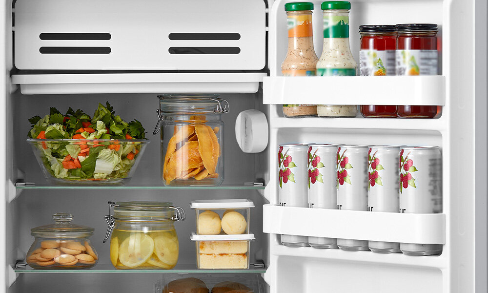 LODÓWKA MIDEA MDRD142FGE50 Zdjęcie przedstawia wnętrze lodówki wypełnione świeżą żywnością, taką jak sałatka, owoce cytrusowe, przekąski i napoje w puszkach. Na drzwiach lodówki umieszczono butelki z sosami oraz soki lub przetwory w szklanych butelkach praktycze wymiary pojemność 93 litry 86 x 47,2 x 45 cm kompaktowa
