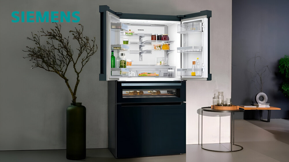 LODÓWKA SIEMENS KF96RSBEA Chłodziarko-zamrażarka Siemens z otwartymi drzwiami górnej komory prezentuje jasno oświetlone wnętrze z półkami i pojemnikami. Urządzenie ustawione jest w aranżacji salonowo‑kuchennej