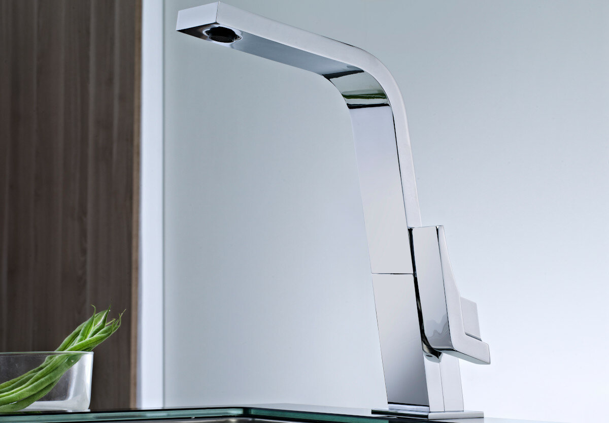 Na obrazie widoczna jest nowoczesna, chromowana bateria umywalkowa o prostokątnym kształcie. W tle znajduje się fragment drewnianej ściany oraz szklana powierzchnia z zieloną rośliną.