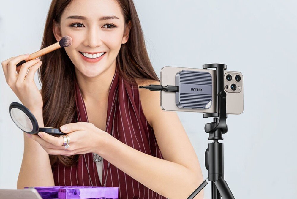 Obudowa dysku UNITEK S1244AGY01 Uśmiechnięta kobieta wykonująca makijaż przed smartfonem zamocowanym na statywie. Do tylnej obudowy telefonu przyczepiony jest dysk Unitek połączony z nim krótkim przewodem