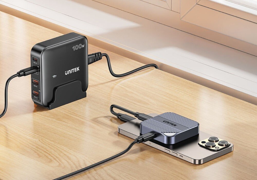 Obudowa dysku UNITEK S1244AGY01 Smartfon leżący na drewnianym stole z podpiętym dyskiem Unitek. Całość jest jednocześnie ładowana przez kabel biegnący od czarnej ładowarki sieciowej Unitek o mocy 100W stojącej obok