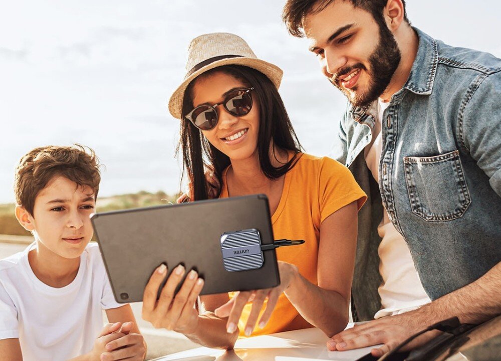 Obudowa dysku UNITEK S1244AGY01 Grupa trzech osób na zewnątrz spoglądająca na tablet trzymany przez kobietę. Do tylnej części obudowy tabletu przylega małe, kwadratowe urządzenie marki Unitek