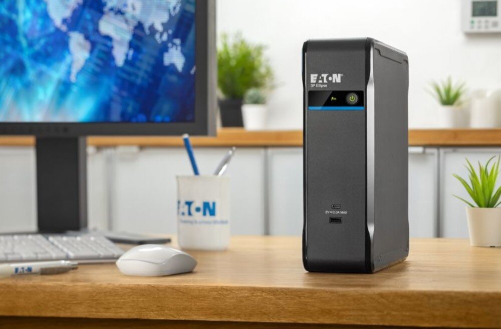 Na biurku znajduje się czarny zasilacz UPS z logo \'EATON\' i napisem \'3P Ellipse\'. W tle widoczny jest monitor, myszka, kubek z długopisami oraz rośliny doniczkowe.