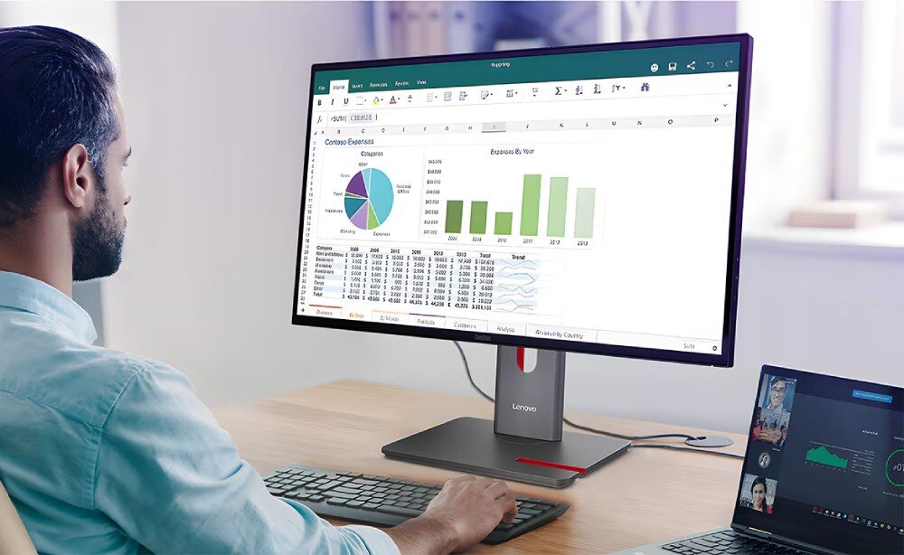 Monitor LENOVO ThinkVision P34WD-40 Widok zza ramienia mężczyzny na monitor wyświetlający arkusz kalkulacyjny z kolorowymi wykresami kołowymi i słupkowymi oraz tabelami danych, obok na blacie stoi otwarty laptop z widokiem uczestników wideokonferencji, odświeżanie, matryca, płynność