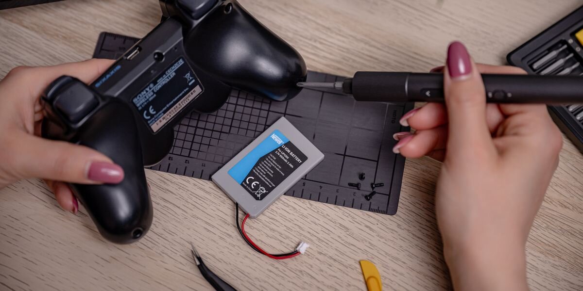 Akumulator NEWELL 650 mAh LIP1359 do kontrolera DualShock 3 Sony PlayStation 3, Dłonie przykręcające obudowę czarnego kontrolera Sixaxis śrubokrętem. Obok leży wymieniony akumulator, przeładowanie, przegrzanie, zwarcie, IC