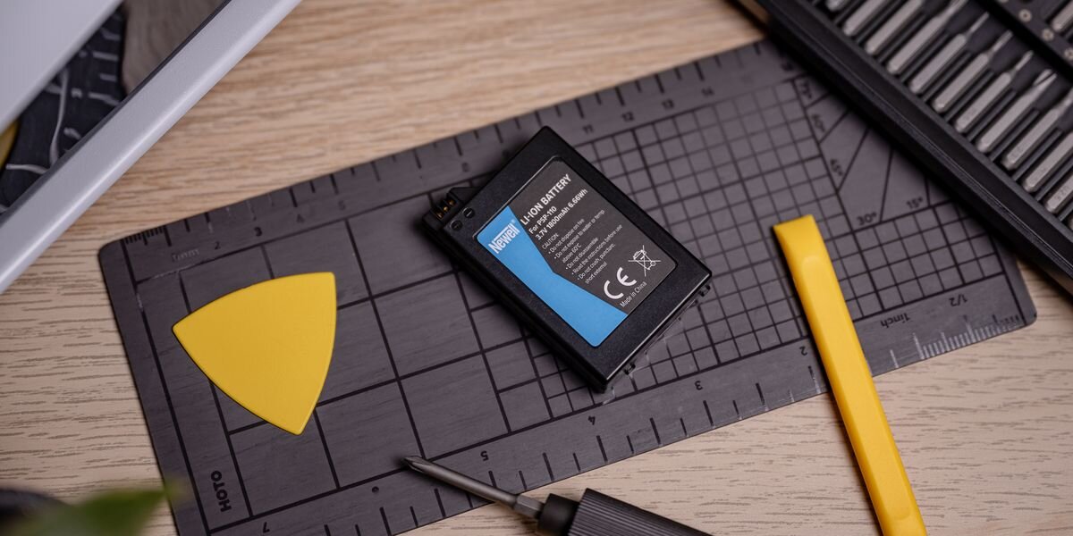 Na stole leży akumulator z napisem \'NEWELL Li-ion BATTERY\', obok niego znajduje się żółty trójkątny element oraz czarny śrubokręt. W tle widoczna jest czarna mata z miarką i żółty plastikowy narzędzie.