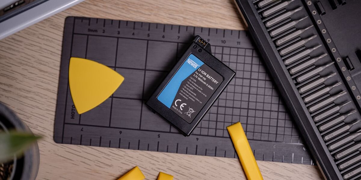 Na zdjęciu znajduje się akumulator z widocznym logo \'Newell\' oraz tekstem \'Li-Ion Battery\'. Obok akumulatora leżą żółte narzędzia na czarnej macie z miarką.