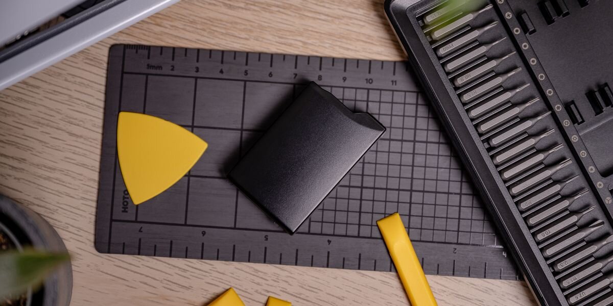 Na drewnianym blacie leży czarny akumulator na czarnej macie z miarką. Obok znajdują się żółte narzędzia i zestaw końcówek w etui.