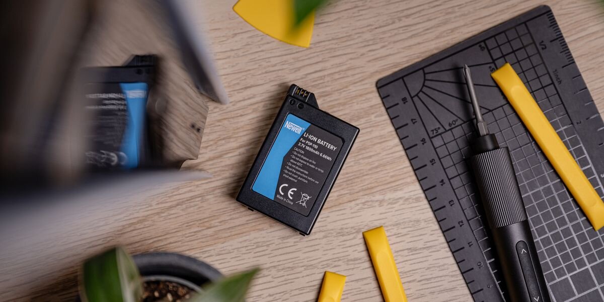 Na drewnianym blacie leży akumulator z widocznym napisem \'NEWELL Li-ion Battery 1800 mAh 3.6V 6.5Wh\'. Obok znajdują się narzędzia i żółte elementy plastikowe.