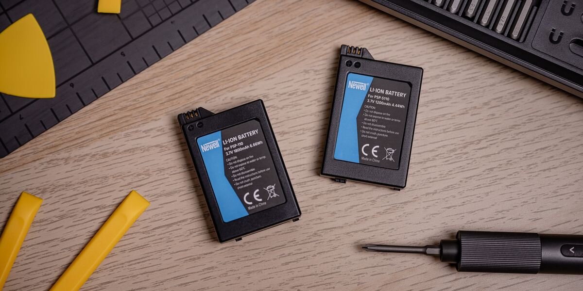 Na drewnianym blacie leżą dwa akumulatory z napisem \'NEWELL Li-Ion Battery 3.7V 1800mAh 6.66Wh\', obok nich znajduje się śrubokręt i żółte narzędzia plastikowe. W tle widoczna jest czarna mata z miarką.