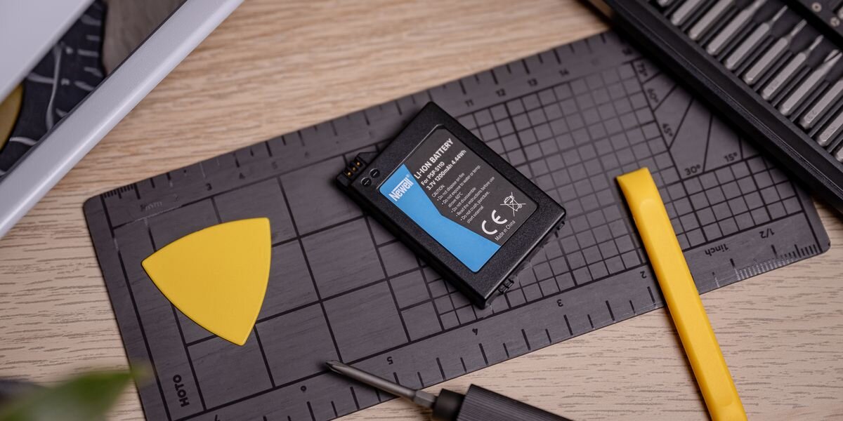 Akumulator NEWELL 1200 mAh PSP-S110 do Sony PlayStation Portable PSP-2000 / PSP-3000, Czarna bateria Newell do PSP na szarej macie serwisowej obok żółtej kostki do podważania obudowy i wkrętaka, energia, PSP, pad, zasilanie