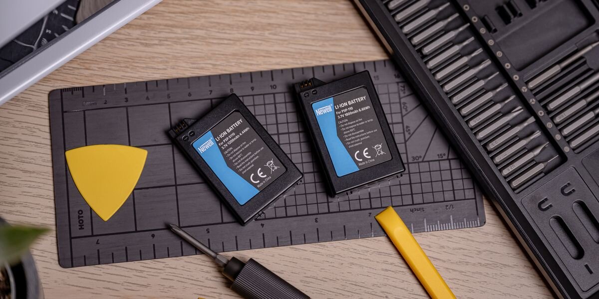 Akumulator NEWELL 1200 mAh PSP-S110 do Sony PlayStation Portable PSP-2000 / PSP-3000, Dwie czarne baterie Newell o różnych pojemnościach na macie serwisowej obok rozbudowanego zestawu bitów, przeładowanie, przegrzanie, zwarcie, Li-ion