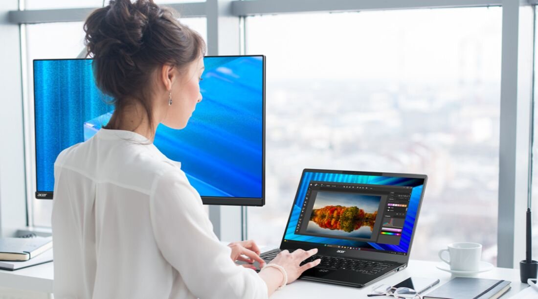 Kobieta w białej koszuli pracująca na laptopie Acer TravelMate z edycją zdjęć w jasnym biurze z zewnętrznym monitorem Acer białą filiżanką kawy notebookami i tabletem graficznym w nowoczesnym wnętrzu z dużymi oknami -  PurifiedView 2.0 z NPU  