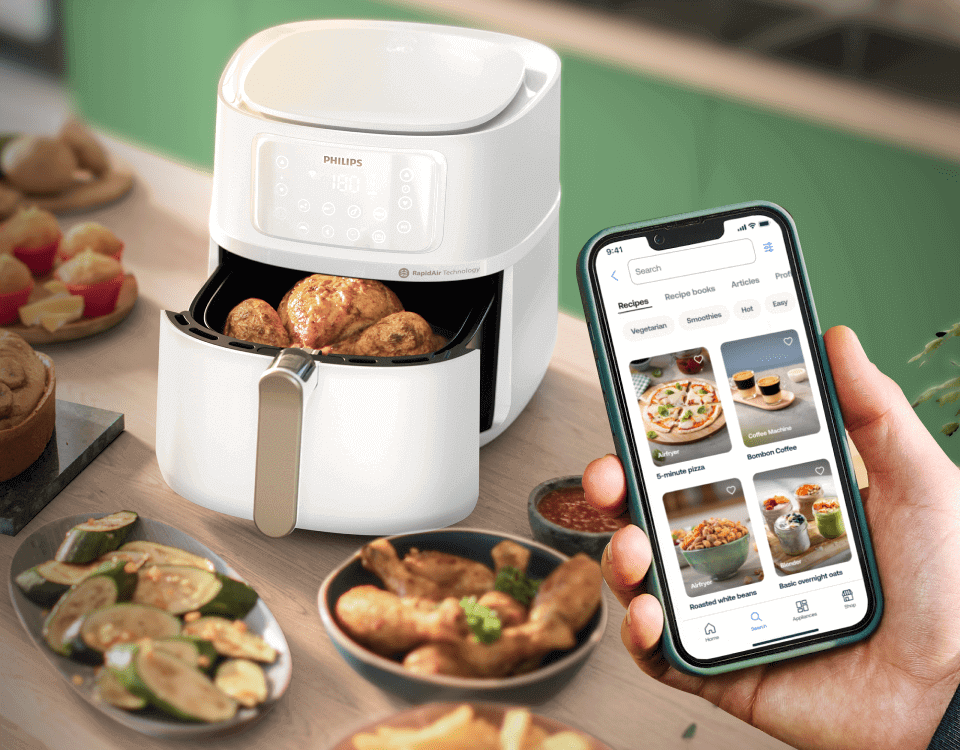 Na blacie kuchennym stoi biała frytkownica beztłuszczowa z logo PHILIPS, z otwartą szufladą pełną ziemniaków. Obok osoba trzyma smartfon z aplikacją pokazującą przepisy kulinarne.