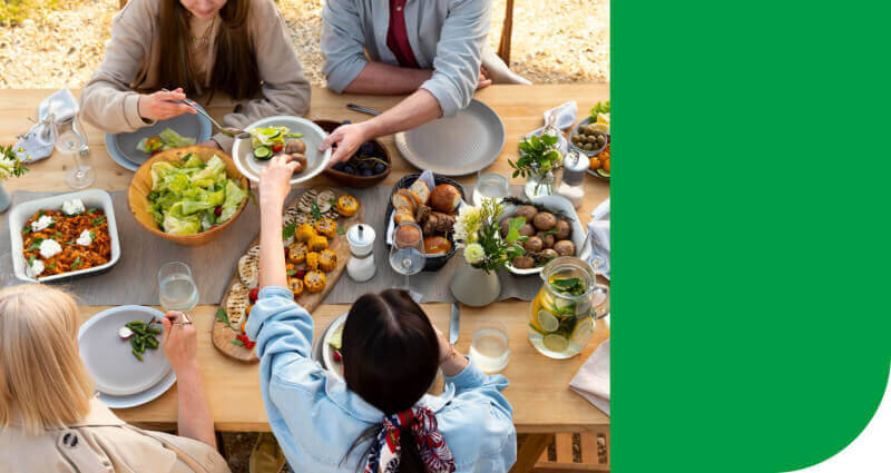 Grupa osób siedzi przy drewnianym stole zastawionym różnorodnymi potrawami, w tym sałatką, pieczywem i napojami. Obok znajduje się zielony prostokąt wypełniający część obrazu.