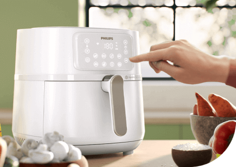 Biała frytkownica beztłuszczowa z panelem dotykowym, na którym widnieje logo PHILIPS i ustawiona temperatura 180°C. Obok frytkownicy znajduje się ręka dotykająca panel oraz miska z warzywami.