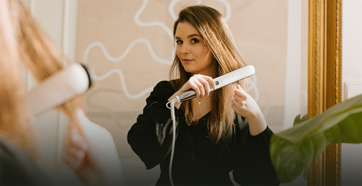 Osoba używa prostownicy do włosów w jasnym pomieszczeniu. Prostownica jest biała i ma widoczne przyciski na uchwycie.