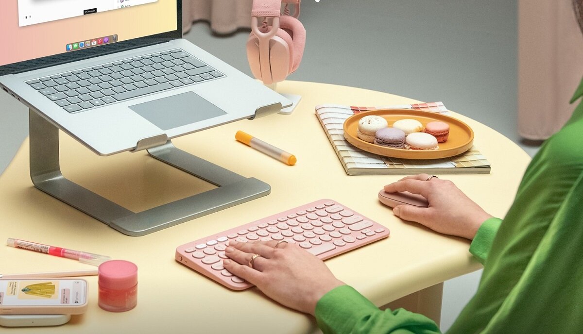 Na biurku znajduje się różowa klawiatura i myszka, obok laptopa na stojaku. W tle widoczne są słuchawki, talerz z makaronikami i kilka przyborów biurowych.