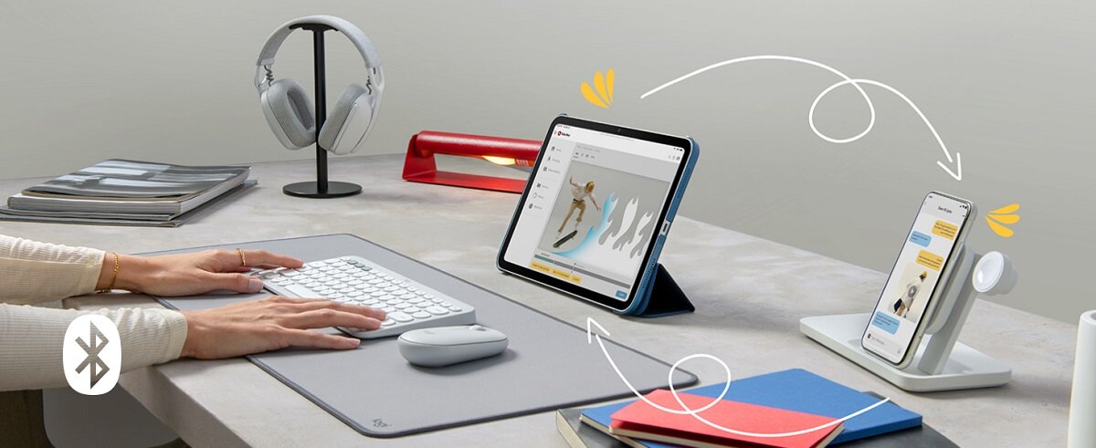 Na biurku znajduje się klawiatura i myszka, obok stoją tablet i smartfon na stojakach. W tle widoczne są słuchawki na stojaku oraz kilka książek i lampka biurkowa.