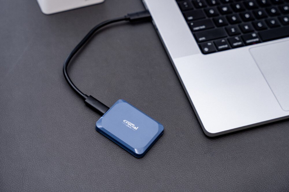 Dysk CRUCIAL X10 Niebieski dysk podłączony kablem USB-C do srebrnego laptopa leżącego na ciemnym blacie, wydajność, szybkość, interfejs, złącze