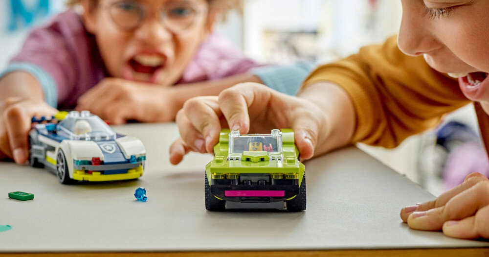 Dwoje dzieci bawi się dwoma samochodami z klocków LEGO na stole. Jeden samochód jest zielony, a drugi biało-niebieski z emblematem przypominającym policyjny.
