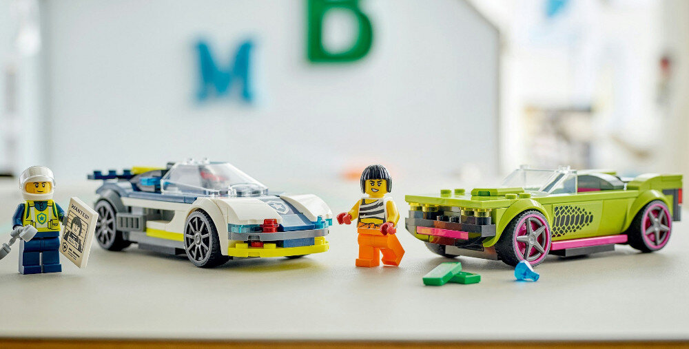 Na obrazie znajdują się dwa kolorowe samochody z klocków LEGO oraz dwie figurki postaci. W tle widoczne są rozmyte litery M i B.