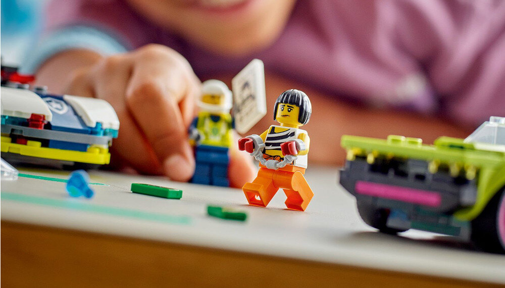 Na obrazie widać dwie figurki LEGO, z których jedna trzyma tabliczkę, oraz fragmenty pojazdów LEGO. W tle widoczna jest ręka osoby bawiącej się zestawem.