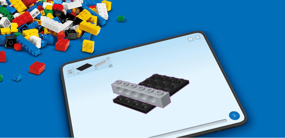 Na niebieskim tle znajduje się tablet z wyświetlonym obrazem dwóch klocków LEGO, jeden szary, drugi czarny. Obok tabletu leży stos kolorowych klocków LEGO.
