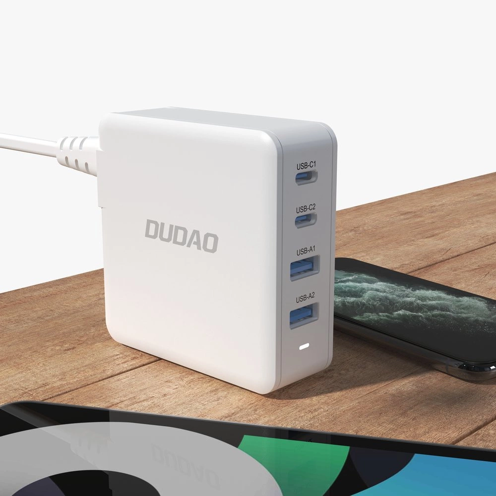 Biała ładowarka sieciowa z logo \'DUDAO\' posiada cztery porty: dwa USB-C oznaczone jako USB-C1 i USB-C2 oraz dwa USB-A oznaczone jako USB-A1 i USB-A2. Obok ładowarki leży smartfon, a w tle widać fragment innego urządzenia z kolorowym ekranem.