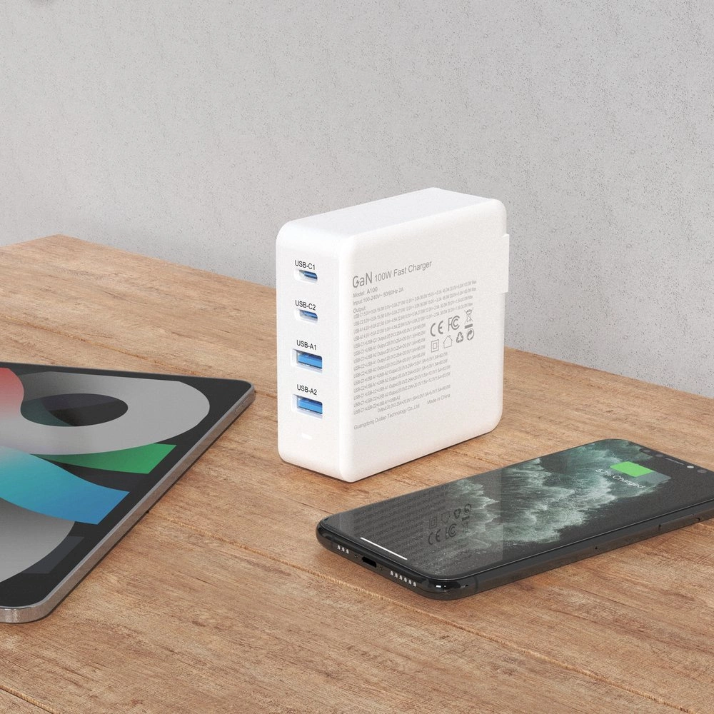 Na drewnianym blacie znajduje się biała ładowarka sieciowa z czterema portami USB oznaczonymi jako USB-C1, USB-C2, USB-A1, USB-A2. Obok leży smartfon z zieloną ikoną baterii oraz tablet z kolorowym ekranem.