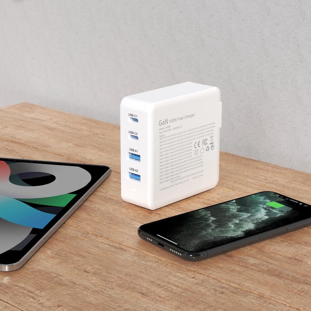 Na drewnianym blacie stoi biała szybka ładowarka sieciowa z czterema portami USB oznaczonymi jako USB-C1, USB-C2, USB-A1, USB-A2. Obok ładowarki leży tablet z kolorowym ekranem oraz smartfon z wyświetlonym wskaźnikiem ładowania.