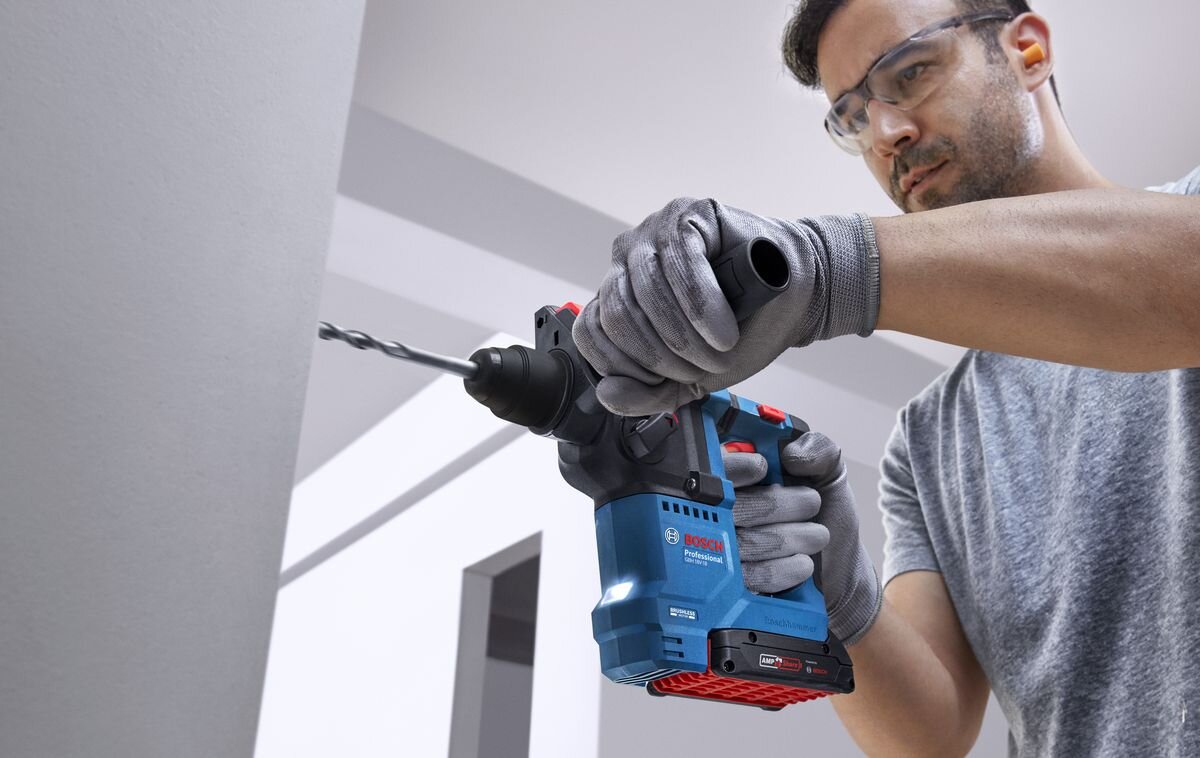 BOSCH Professional GBH 18V-18 0611927001 Funkcja udaru nominalnej energii udaru 1,5 J liczbie udarów do 5000 obr/min Prędkość obrotowa bez obciążenia wynosi 0–900 obr/min - Mężczyzna w okularach ochronnych trzyma oburącz pracujący młot udarowo-obrotowegy przy ścianie.