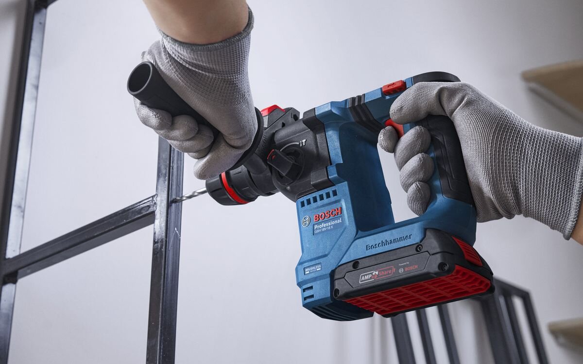 Osoba w rękawicach trzyma niebieski młot udarowo-obrotowy z widocznym logo BOSCH Professional. Narzędzie jest używane do wiercenia w metalowej konstrukcji.