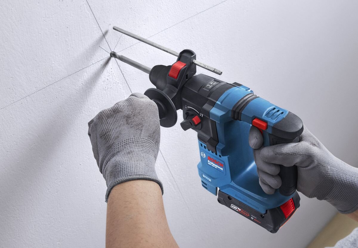 BOSCH Professional GBH 18V-18 0611927002 BOSCH Professional GBH 18V-18 0611927002 Auto Power GDE 12 bezpyłową pracę młotami bez odsysania pyłu - Mężczyzna w okularach ochronnych obsługuje urządzenie przy ścianie z zaznaczonymi punktami wiercenia.