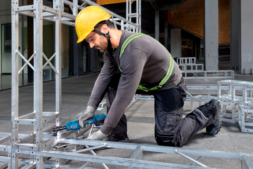 Klucz akumulatorowy grzechotka BOSCH GRC 18V-60 06019N8001 pomieszczenie plac budowy osoba skręca metalowe elementy w tle okna szklane drzwi obsługuje standardowe śruby w zakresie M5–M12 do szerokiego zakresu zastosowań w warsztacie, serwisie samochodowym i montażu konstrukcji stalowych uchwyt 3/8 cala szybkie mocowanie nasadek stabilność pracy ogranicza ryzyko błędów i konieczności poprawiania wykonanych połączeń