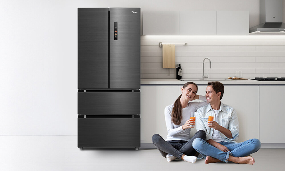 Na obrazie widoczna jest antracytowa lodówka z dwoma górnymi drzwiami i dwoma dolnymi szufladami. Obok lodówki siedzą dwie osoby na podłodze, trzymając napoje.