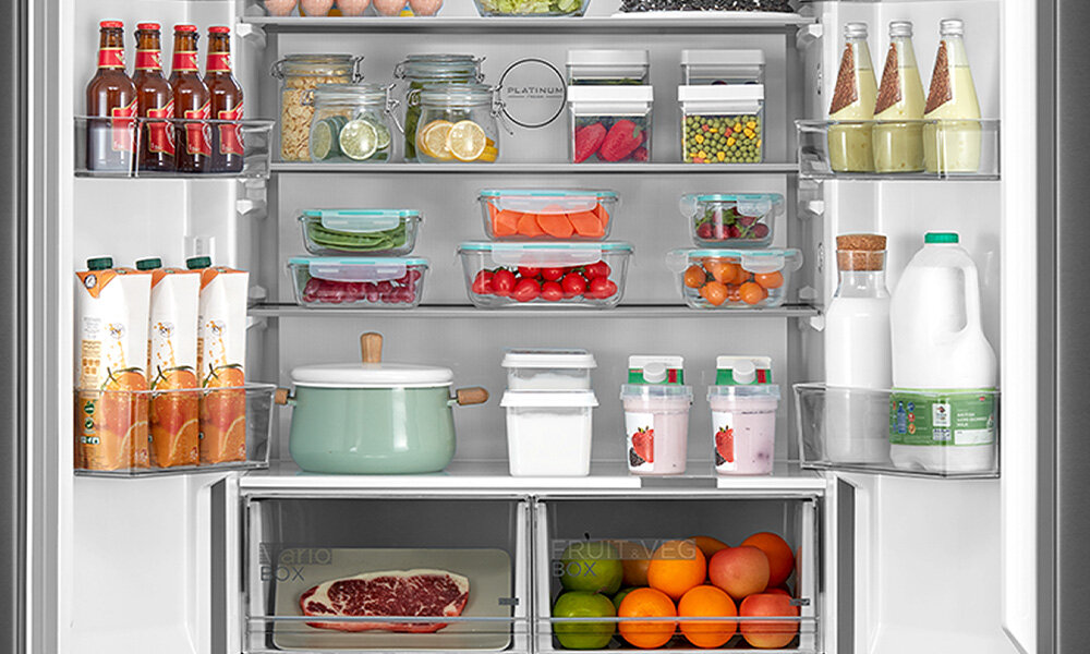 Wnętrze lodówki wypełnione różnorodnymi produktami spożywczymi, w tym napojami, słoikami z przetworami, warzywami i owocami. Na drzwiach widoczne są butelki i kartony z napojami, a na półkach wewnętrznych pojemniki z jedzeniem oraz garnki.